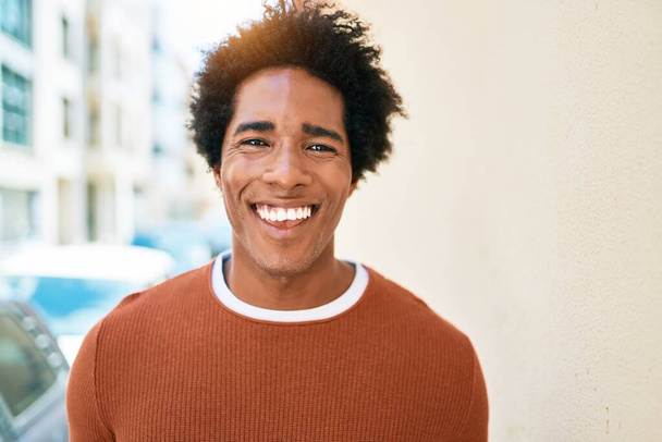 Νεαρός όμορφος Αφροαμερικάνος που φοράει καθημερινά ρούχα χαμογελώντας χαρούμενος. Στέκεται με χαμόγελο στο πρόσωπο περπατώντας στο δρόμο της πόλης. - Φωτογραφία, εικόνα