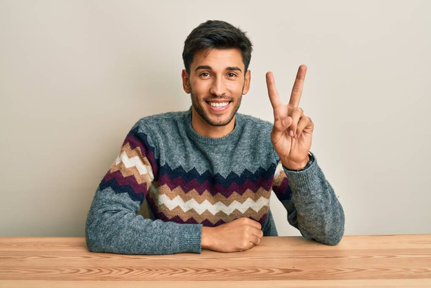 Junger gutaussehender Mann in lässigem Pullover sitzt auf dem Tisch und zeigt mit Finger Nummer zwei nach oben, während er selbstbewusst und glücklich lächelt.  - Foto, Bild