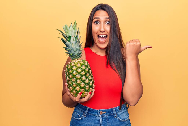 Young beautiful brunette woman holding pineapple pointing thumb up to the side smiling happy with open mouth  - Photo, Image