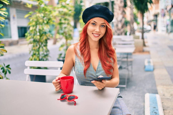 Young redhead girl wearing french style using smartphone sitting at the coffee shop terrace. - Fotó, kép