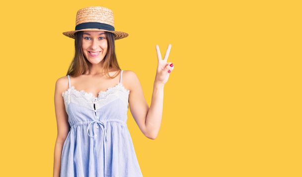 Beautiful brunette young woman wearing summer hat smiling with happy face winking at the camera doing victory sign. number two.  - Photo, Image