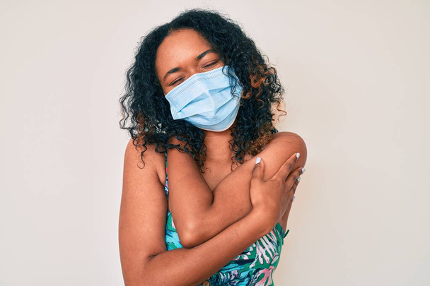 Young african american woman wearing medical mask hugging oneself happy and positive, smiling confident. self love and self care  - Zdjęcie, obraz