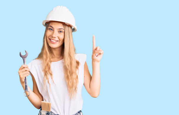 Piękna biała kobieta z blond włosami nosząca twarde kapelusze i ubrania budowlańca zaskoczona pomysłem lub pytaniem wskazującym palcem na szczęśliwą twarz, numer jeden  - Zdjęcie, obraz