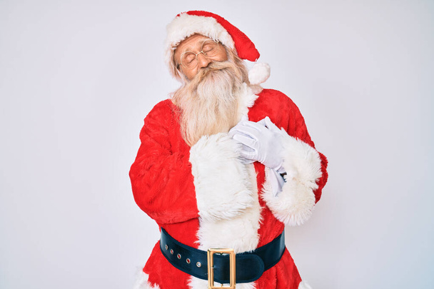 Старик с седыми волосами и длинной бородой в костюме Санта Клауса с подтяжками, улыбающимися руками на груди с закрытыми глазами и благодарным жестом на лице. концепция здоровья.  - Фото, изображение