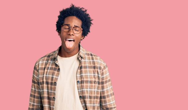 カジュアルな服を着たアフロの髪をしたハンサムなアフリカ系アメリカ人男性と面白い表情で幸せ舌を突き出す眼鏡。感情の概念.  - 写真・画像