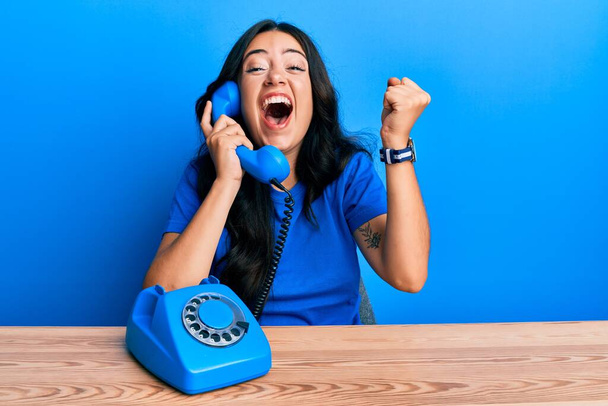 Belle brune jeune femme parlant sur téléphone vintage criant fière, célébrant la victoire et le succès très excitée avec les bras levés  - Photo, image