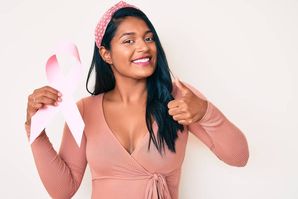 Schöne junge Frau mit langen Haaren, die ein rosafarbenes Krebsband in der Hand hält und glücklich und positiv lächelt, Daumen hoch und Zeichen der Anerkennung  - Foto, Bild
