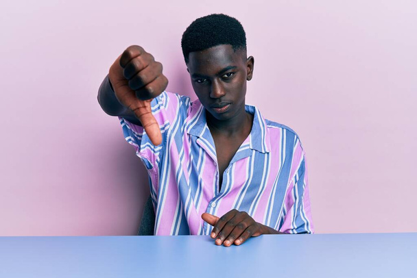 Young african american man wearing casual clothes sitting on the table looking unhappy and angry showing rejection and negative with thumbs down gesture. bad expression.  - Photo, Image