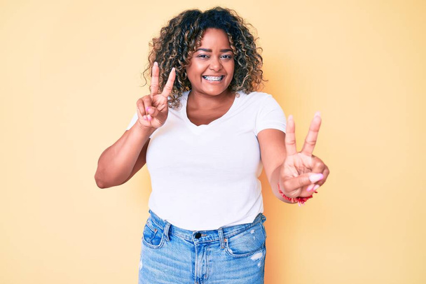 Jonge Afro-Amerikaanse plus size vrouw draagt casual wit tshirt glimlachend naar de camera kijkend met vingers die een overwinningsteken doen. nummer twee.  - Foto, afbeelding