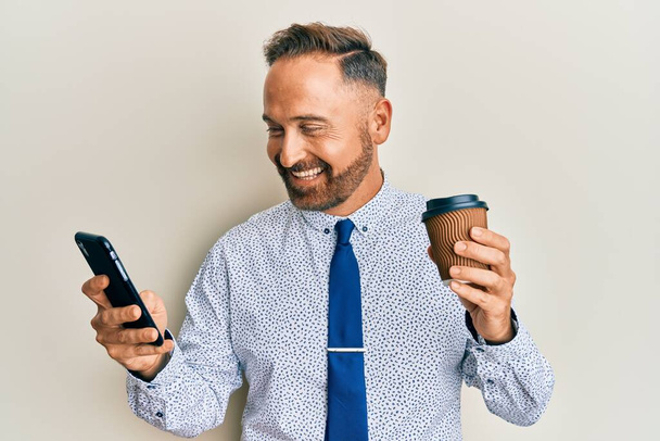 Hombre de negocios guapo de mediana edad usando un teléfono inteligente y bebiendo una taza de café sonriendo y riendo en voz alta porque broma loca divertida.  - Foto, imagen