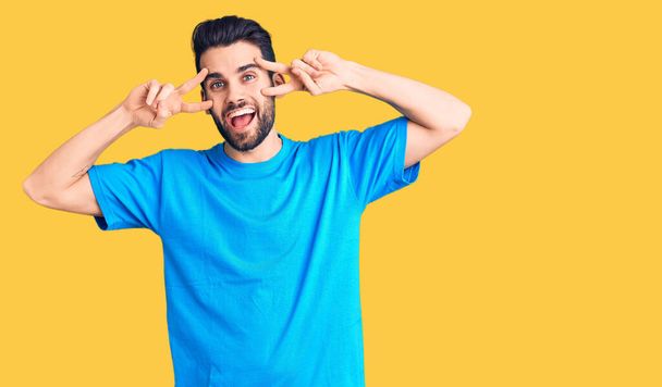 Young handsome man with beard wearing casual t-shirt doing peace symbol with fingers over face, smiling cheerful showing victory  - Foto, Bild