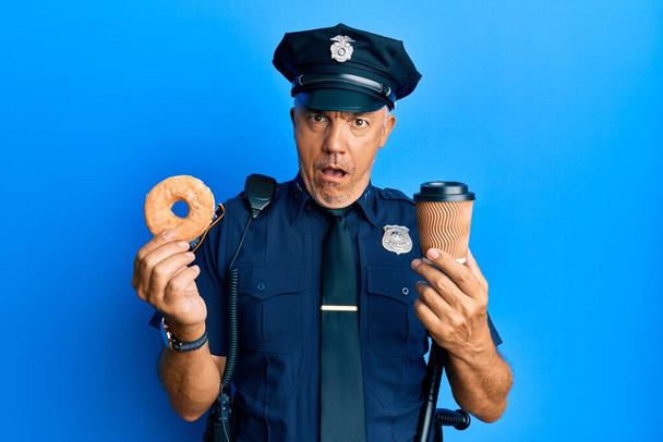 Fešák středního věku zralý policista jíst koblihy a pití kávy v šoku tvář, vypadající skeptický a sarkastický, překvapený s otevřenými ústy  - Fotografie, Obrázek