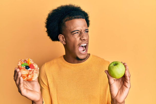African american man with afro hair holding pastry and healthy green apple angry and mad screaming frustrated and furious, shouting with anger. rage and aggressive concept.  - Foto, Imagen