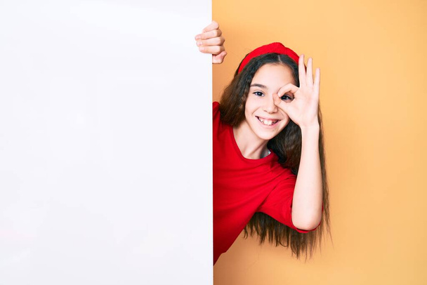 Mignon hispanique enfant fille tenant vide bannière souriante heureux faire ok signe avec la main sur l'œil regardant à travers les doigts  - Photo, image