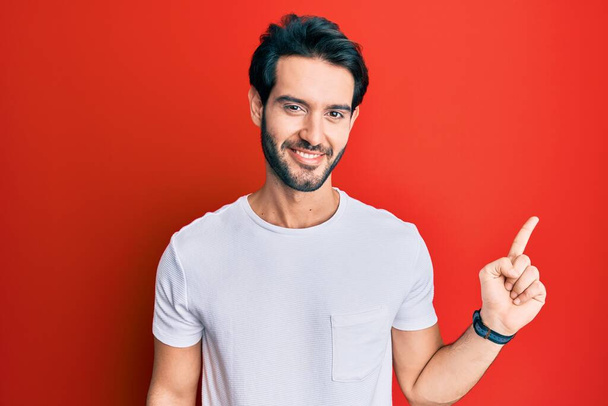 Joven hispano vistiendo una camiseta blanca casual con una gran sonrisa en la cara, señalando con el dedo de la mano hacia un lado mirando a la cámara.  - Foto, Imagen