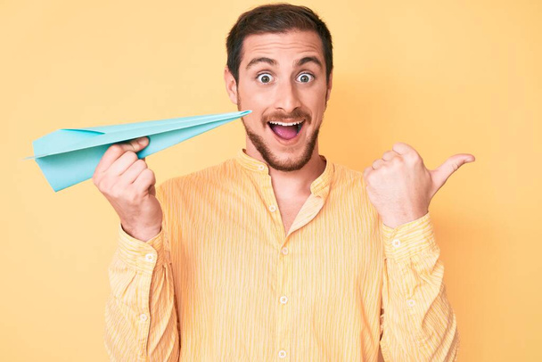 Young handsome man holding paper airplane pointing thumb up to the side smiling happy with open mouth  - Foto, imagen