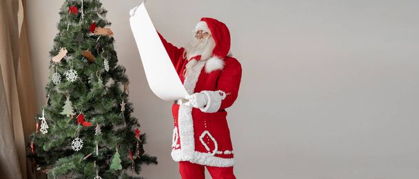 Święty Mikołaj stoi obok choinki trzymając kartkę papieru w rękach. Koncepcja dobrej listy dzieci - Zdjęcie, obraz