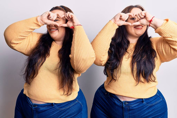 Young plus size twins wearing casual clothes doing heart shape with hand and fingers smiling looking through sign  - Foto, afbeelding