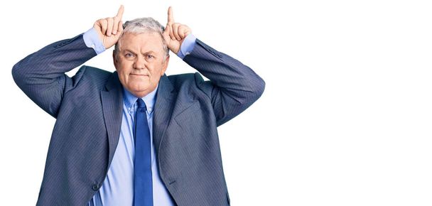 Hombre de pelo gris senior con chaqueta de negocios haciendo un gesto divertido con el dedo sobre la cabeza como cuernos de toro  - Foto, imagen
