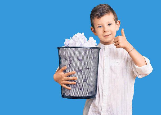 Χαριτωμένο ξανθό παιδί κρατώντας χάρτινο κάδο γεμάτο τσαλακωμένα χαρτιά χαμογελώντας χαρούμενος και θετικός, ο αντίχειρας επάνω κάνει εξαιρετική και την έγκριση σημάδι  - Φωτογραφία, εικόνα