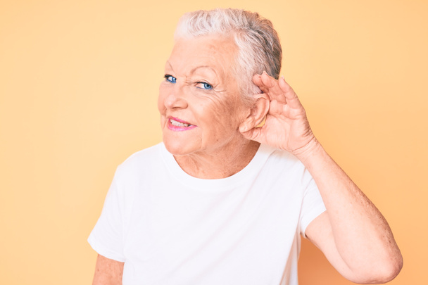 Belle femme âgée aux yeux bleus et aux cheveux gris portant un t-shirt blanc classique sur fond jaune souriant avec main sur oreille en écoutant une rumeur ou des commérages. concept de surdité.  - Photo, image