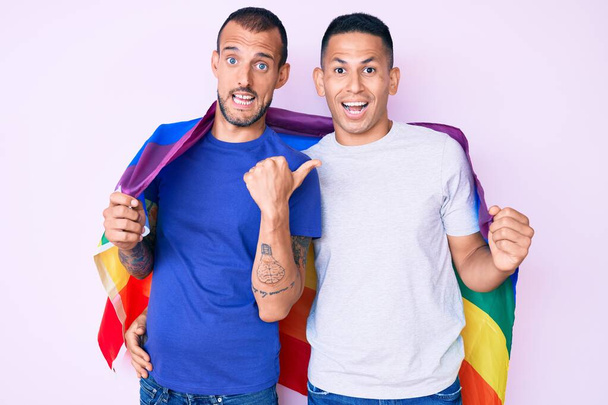 Young gay couple of two men holding rainbow lgbtq flag together pointing thumb up to the side smiling happy with open mouth  - Foto, Imagem