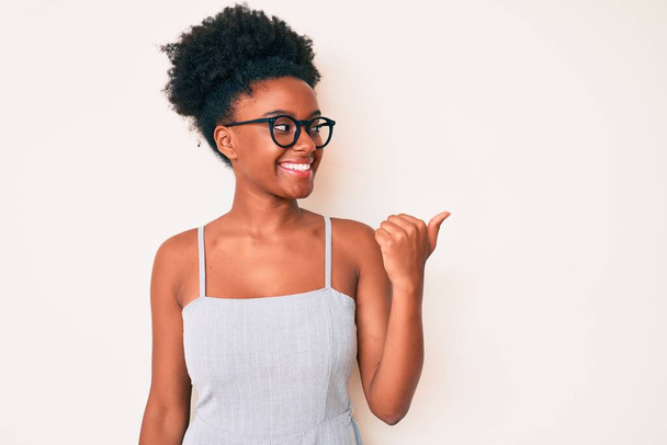 若いアフリカ系アメリカ人女性を着てカジュアルな服と幸せな顔を見て笑顔と親指で側を指してアップ.  - 写真・画像