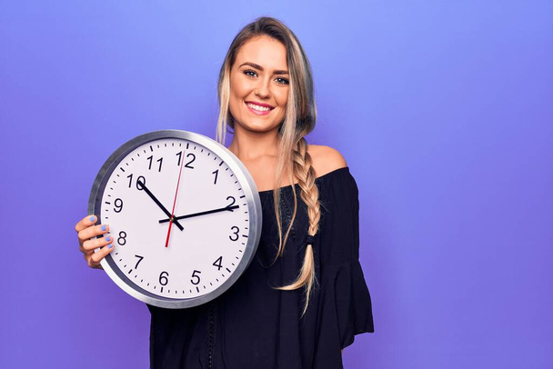 Nuori kaunis blondi nainen tekee lähtölaskenta käyttäen iso kello yli violetti tausta näyttää positiiviselta ja onnellinen seisoo ja hymyilee luottavainen hymy osoittaa hampaat - Valokuva, kuva
