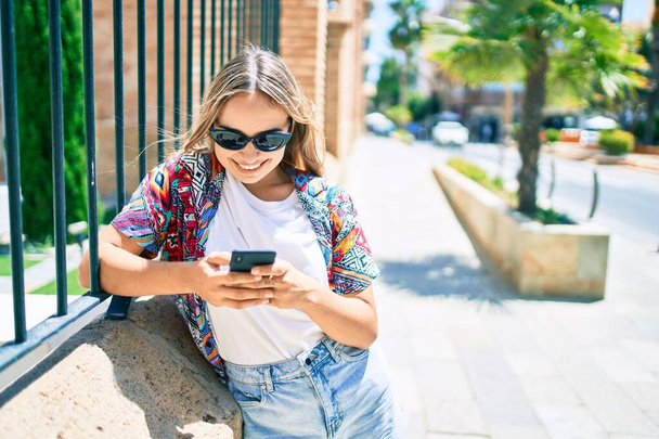 Молодая красивая блондинка-кавказка улыбается на улице в солнечный день, используя смартфон - Фото, изображение