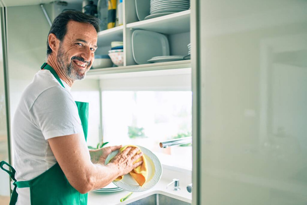 Mężczyzna w średnim wieku z brodą uśmiechnięty szczęśliwy zmywanie naczyń w domu - Zdjęcie, obraz