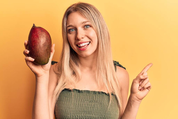 jong mooi blond meisje holding mango glimlachen gelukkig wijzend met hand en vinger naar de kant  - Foto, afbeelding