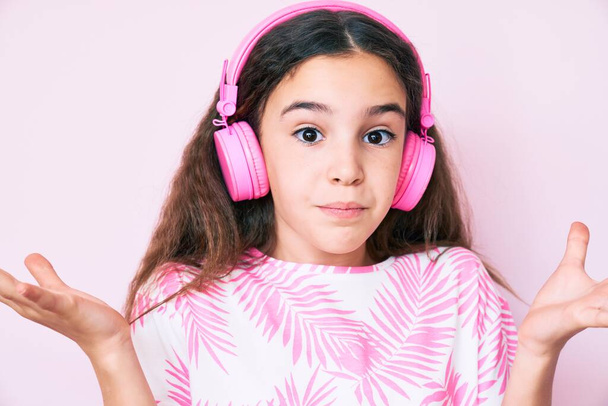 Cute hispanic child girl listening to music using headphones clueless and confused with open arms, no idea and doubtful face.  - Foto, Imagen
