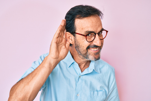 Μεσήλικας Ισπανός που φοράει καθημερινά ρούχα και γυαλιά χαμογελώντας με το χέρι πάνω από το αυτί ακούει και ακούει φήμες ή κουτσομπολιά. έννοια της κώφωσης.  - Φωτογραφία, εικόνα