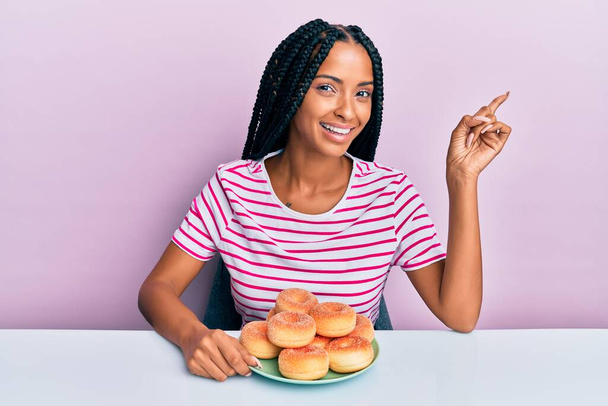 Красива іспаномовна жінка їсть пончик на сніданок, усміхаючись щасливо вказуючи рукою і пальцем в сторону
  - Фото, зображення