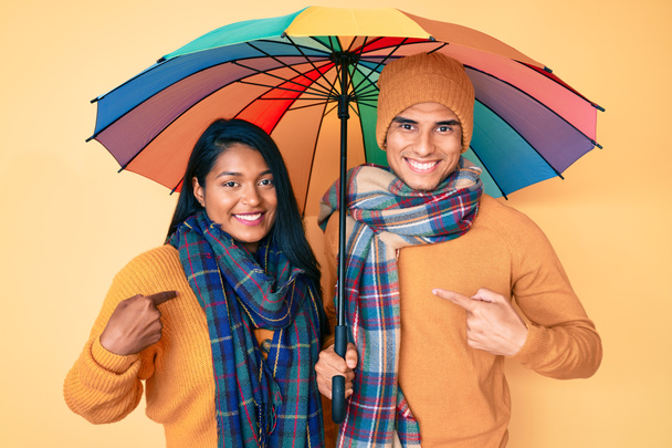 Beautiful latin young couple under colorful umbrella pointing finger to one self smiling happy and proud  - Photo, Image