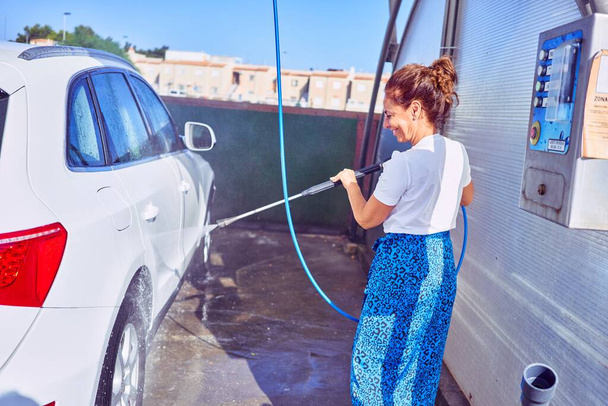 Middle age beautiful woman wearing casual clothes smiling happy. Standing with smile on face washing car using water pistol. - Photo, Image