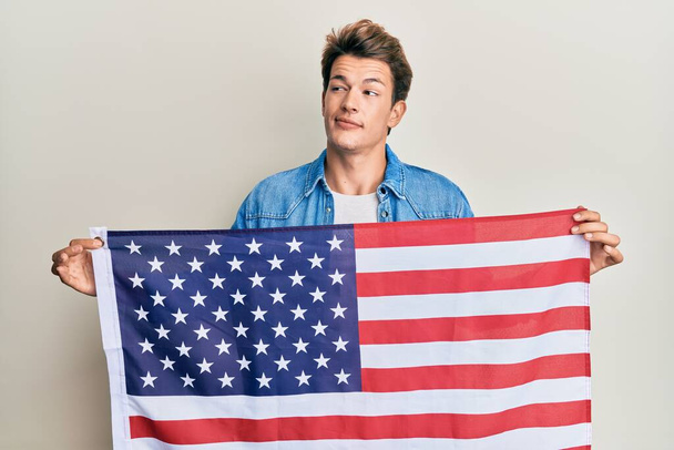bel homme caucasien tenant drapeau des États-Unis souriant regardant vers le côté et regardant loin en pensant.  - Photo, image