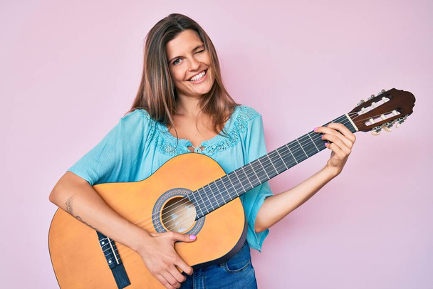 Όμορφη καυκάσια γυναίκα παίζει κλασική κιθάρα κλείνει το μάτι κοιτάζοντας την κάμερα με σέξι έκφραση, χαρούμενο και χαρούμενο πρόσωπο.  - Φωτογραφία, εικόνα