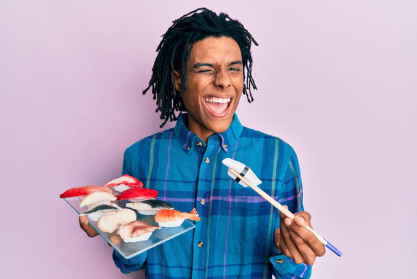 Молодий афроамериканець їсть суші за допомогою паличок, які підморгують, дивлячись на камеру з сексуальним виразом, веселим і щасливим обличчям..  - Фото, зображення