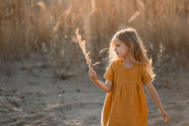 kis szőke lány játszik a folyón a homokban a naplemente ellen. - Fotó, kép