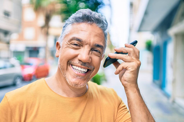 Grauhaariger Mann mittleren Alters lächelt glücklich auf dem Smartphone in der Straße der Stadt. - Foto, Bild