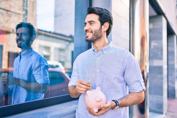 Joven hombre hispano sonriendo feliz ahorro de billetes dentro de alcancía en la ciudad. - Foto, imagen