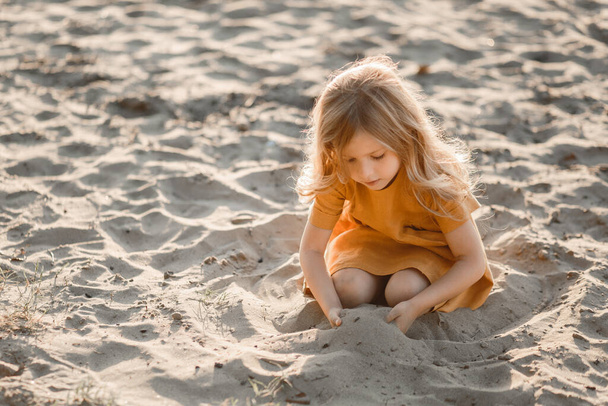 little blonde girl plays on the river in the sand against the sunset sky - Photo, Image