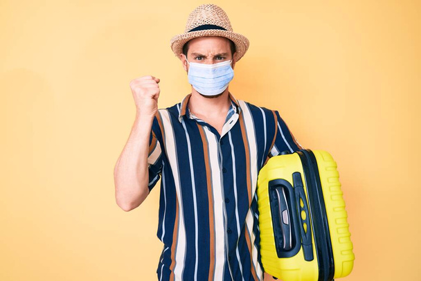 Young handsome man wearing medical mask holding suitcase annoyed and frustrated shouting with anger, yelling crazy with anger and hand raised  - Photo, image