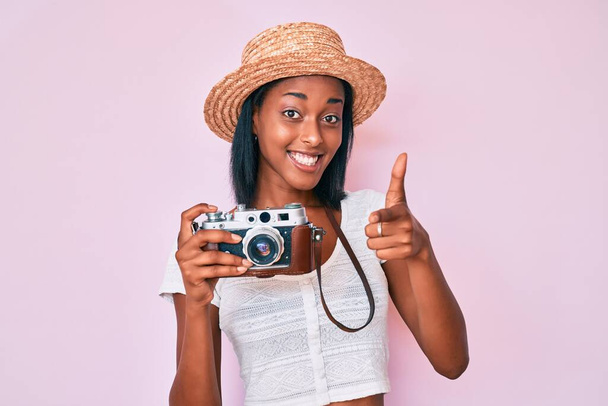 Giovane donna afroamericana con cappello estivo che tiene la fotocamera vintage puntando il dito verso la fotocamera con viso felice e divertente. buona energia e vibrazioni.  - Foto, immagini