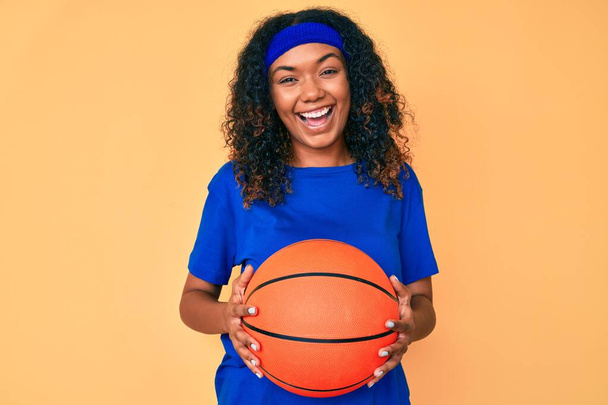 Jeune femme afro-américaine tenant ballon de basket souriant et riant fort à haute voix parce que drôle blague folle.  - Photo, image