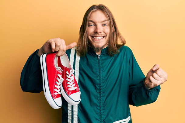 Handsome caucasian man with long hair holding red casual shoes screaming proud, celebrating victory and success very excited with raised arm  - Fotoğraf, Görsel