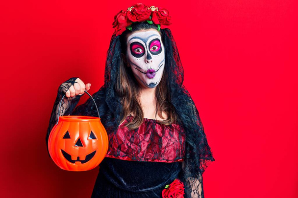 Young woman wearing day of the dead costume holding pumpkin scared and amazed with open mouth for surprise, disbelief face  - Photo, Image