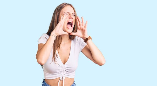 Joven mujer rubia hermosa usando camiseta blanca casual gritando enojado en voz alta con las manos sobre la boca  - Foto, imagen