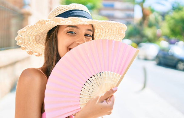 Young hispanic woman on vacation wearing summer style using hand fan at street of city - Photo, Image